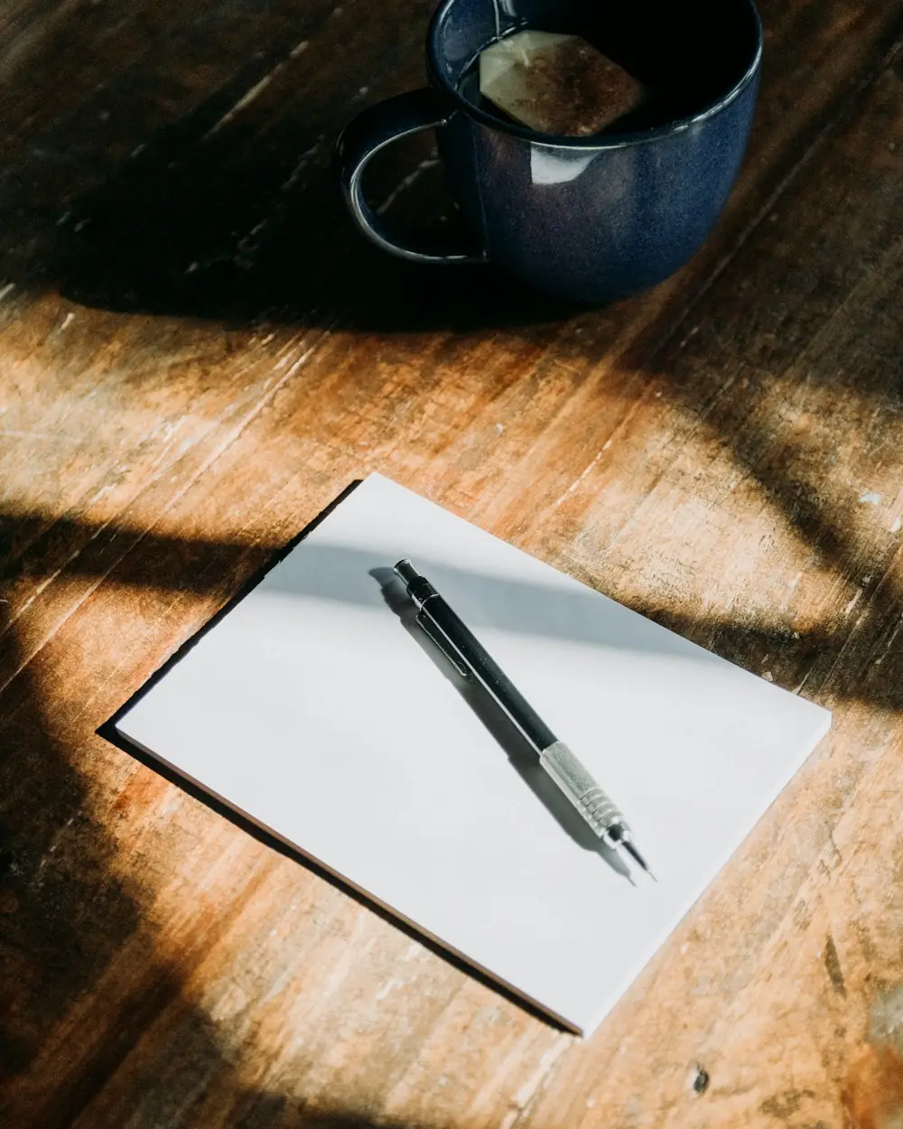 A blank notepad and a mechanical pencil on a sunlit wooden table beside a navy mug of tea.